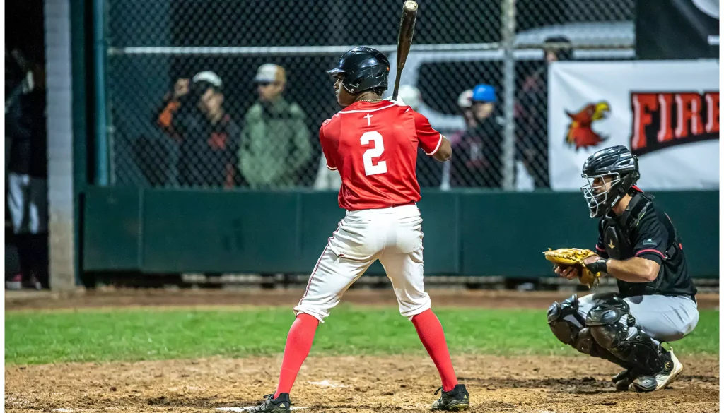 Baseball batter in red jersey number 2 swings a bat, wearing a black helmet, on a dirt field with a catcher nearby in black gear behind him.