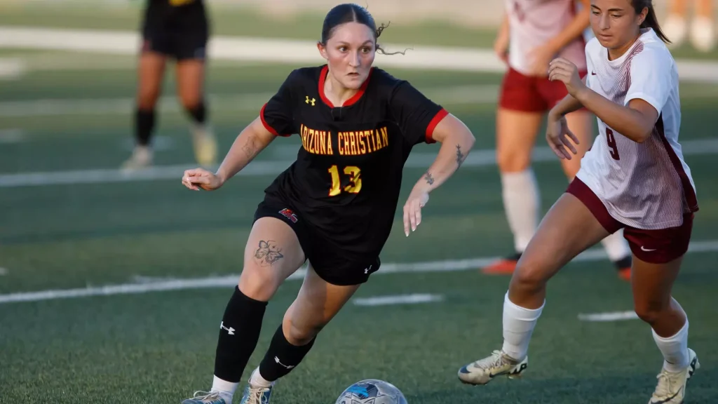 Two female soccer players chase a ball on a grass field during a match; one in a black Arizona Christian jersey (#13) and the other in white/red.