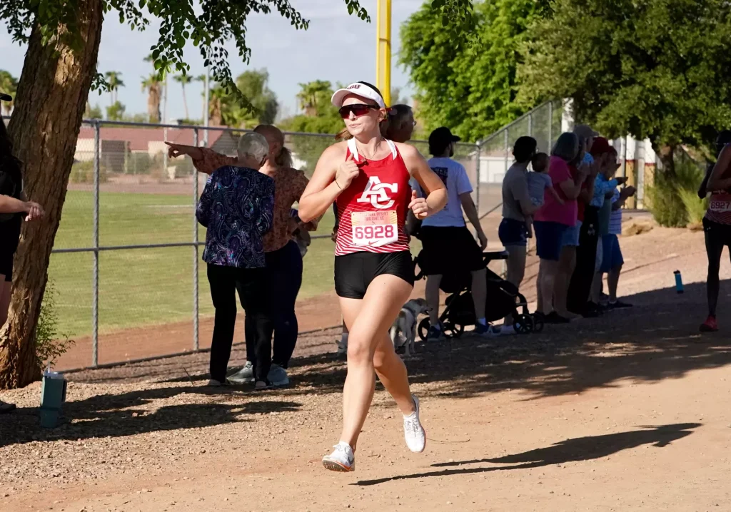 Female runner in a red tank top and white visor, bib number 6928, mid-stride on a dirt path as spectators line a chain-link fence nearby.