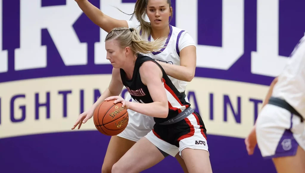 Female basketball players in action: one dribbling the ball while a defender guards closely in a gym with purple signage behind them.