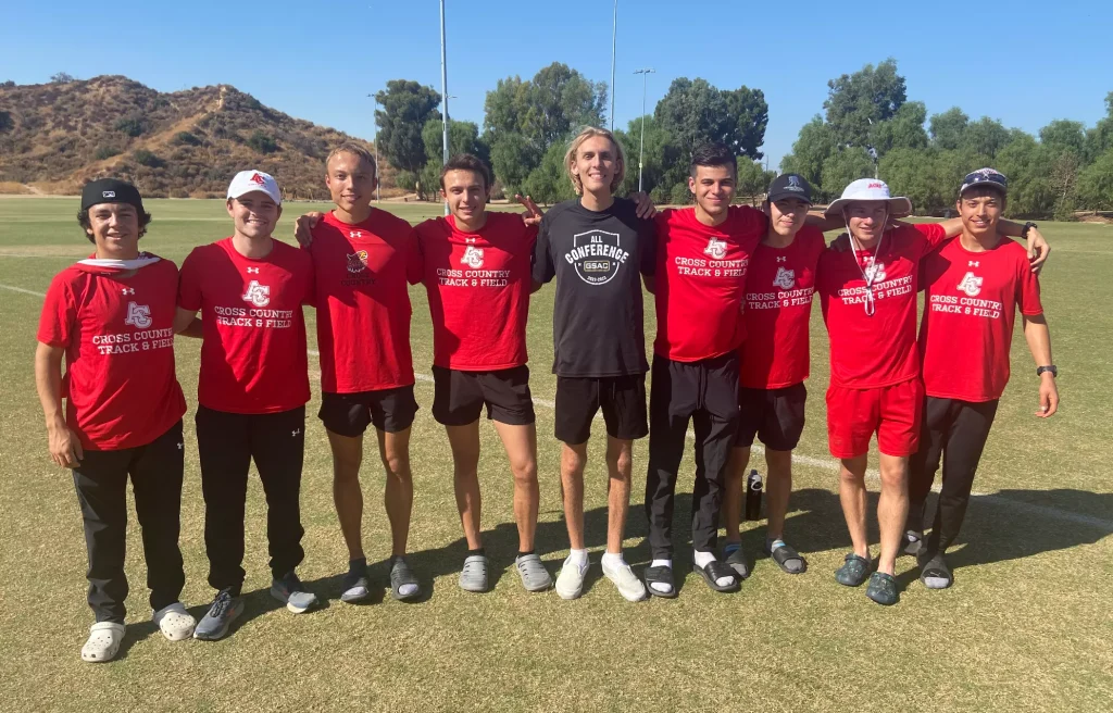 Nine young men in red Cross Country shirts stand shoulder to shoulder on a sunny practice field, arms around each other, smiling with hills in the background.