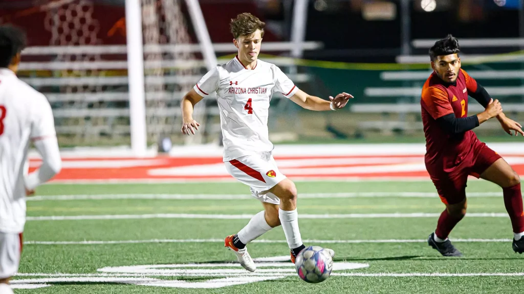 ACU Scores Late Against Park Grabs Last GSAC Playoff Spot - Arizona Christian University - Phoenix, AZ Soccer player in white Arizona Christian kit dribbles the ball on green turf as a red-uniform defender pursues nearby.