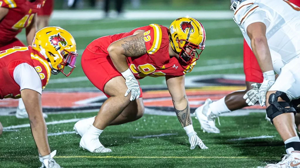 American football players at the line of scrimmage: red uniforms with yellow helmets ready to snap the ball, facing white-uniform defenders.