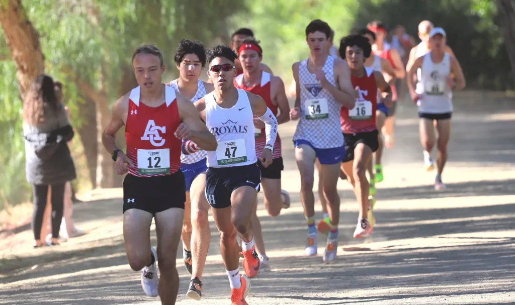 Group of male runners in a road race, sprinting along a tree-lined path; front runner wears a white Royals singlet and bib 47.