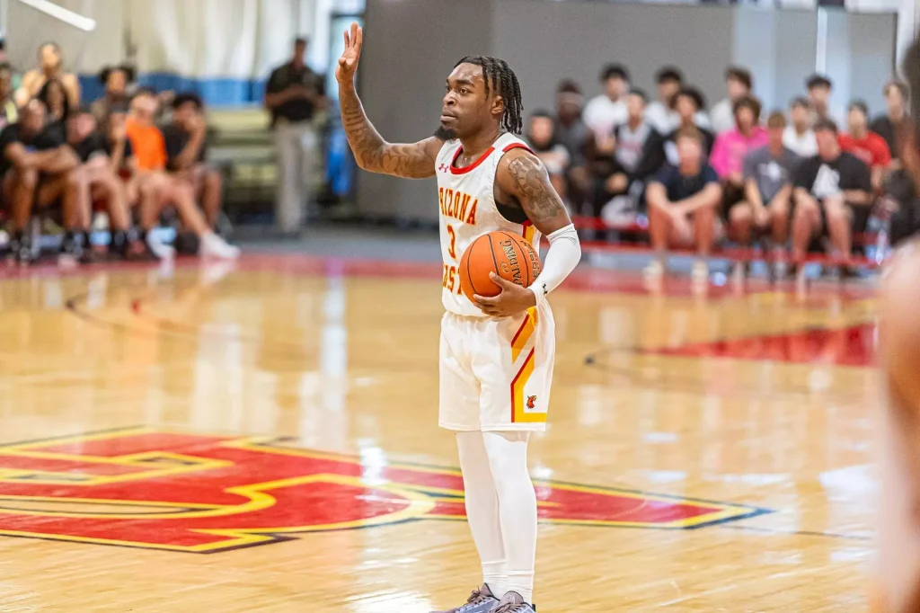 Basketball player in a white Arizona jersey raises his right hand while holding an orange ball on a polished gym floor, with a blurred crowd in the stands behind him.