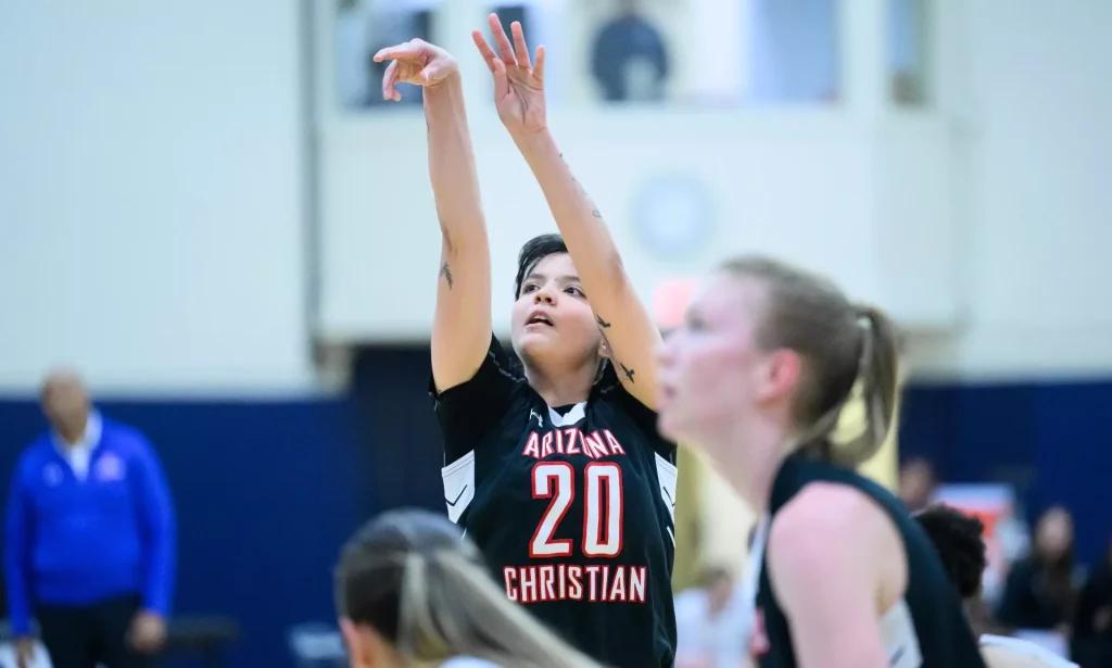 Female basketball player in a black 'Arizona Christian' jersey #20 raises arms for a rebound or shot in a gym, focused on the ball, with teammates in the background.