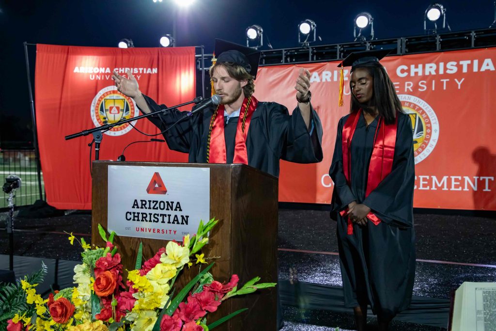Two graduates in black caps and gowns, one speaking at a podium with red tassel, Arizona Christian University backdrop.