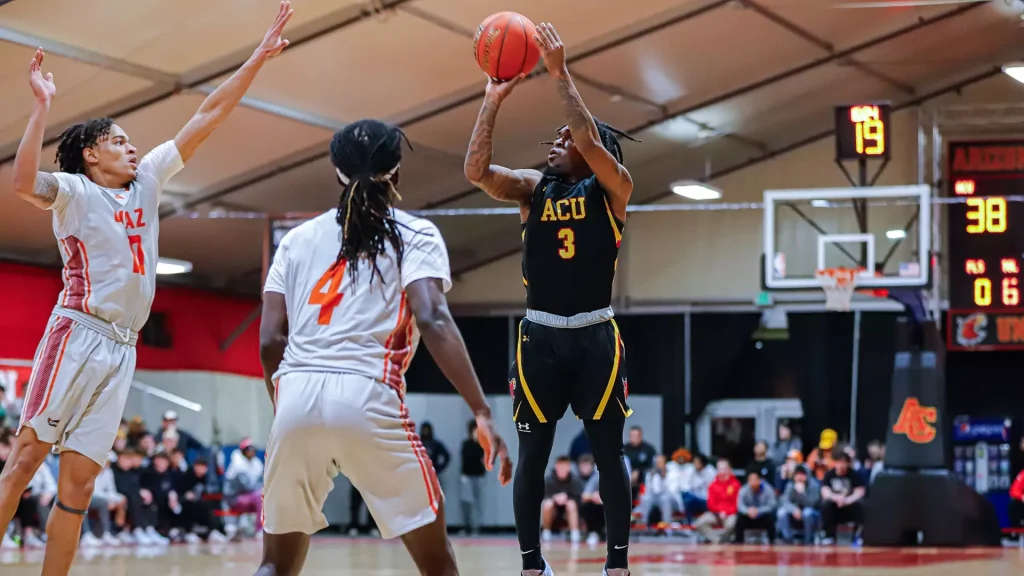 ACU player #3 in black shoots a jump shot while defenders in white react on a crowded indoor court with spectators in the background.