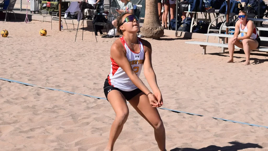 Female beach volleyball player in white and red jersey (#3) prepares to receive a ball on the sandy court, with spectators in the background.