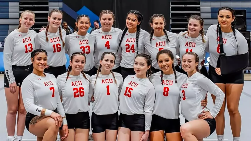ACU STUNT Shows Strong Progress In Early Competitions - Arizona Christian University Group of female athletes in white ACU jerseys posing for a team photo in a gymnasium, smiling at the camera.