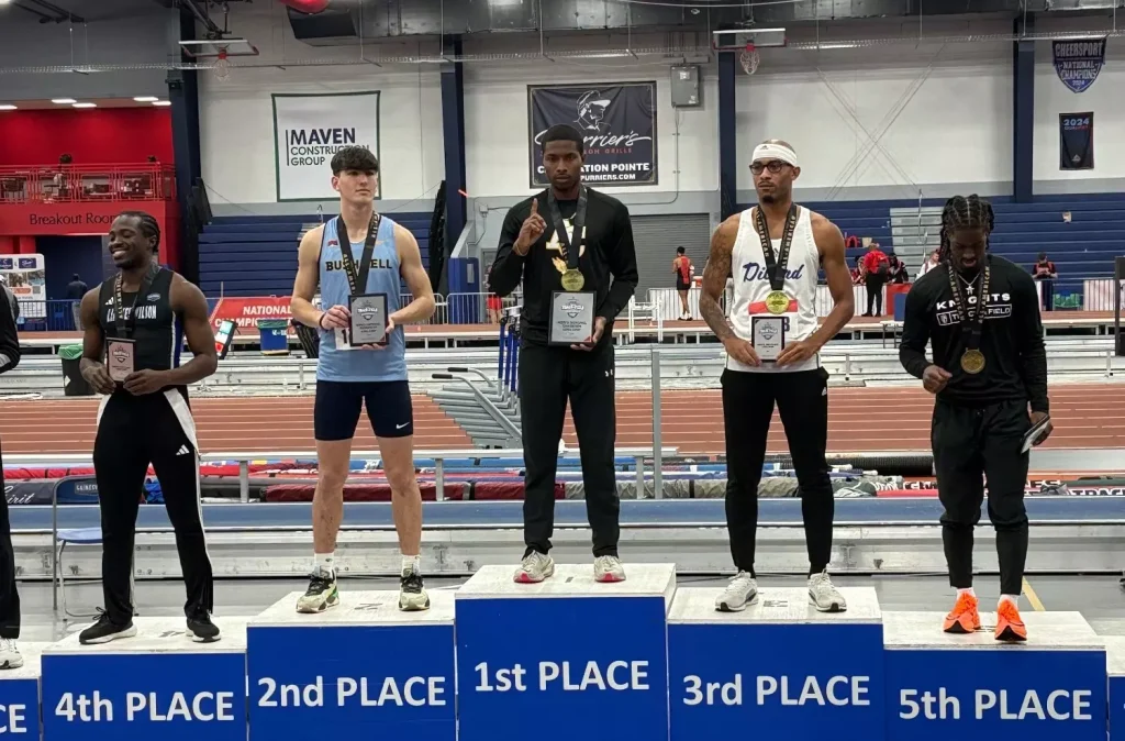Athletes stand on a podium: 1st place center with gold medal and plaque, flanked by 2nd and 3rd place winners, plus two other competitors on the sides in a gymnasium track setting.