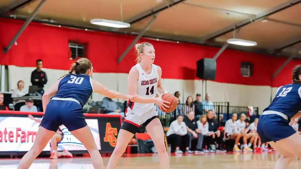 Female basketball player in a white jersey (number 14) dribbles the ball while defended by blue-jersey players in an indoor gym with red walls and spectators watching nearby.