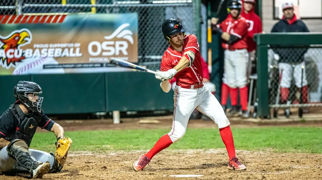 Baseball batter in red uniform swings at a pitch as a catcher crouches behind on the dirt field.