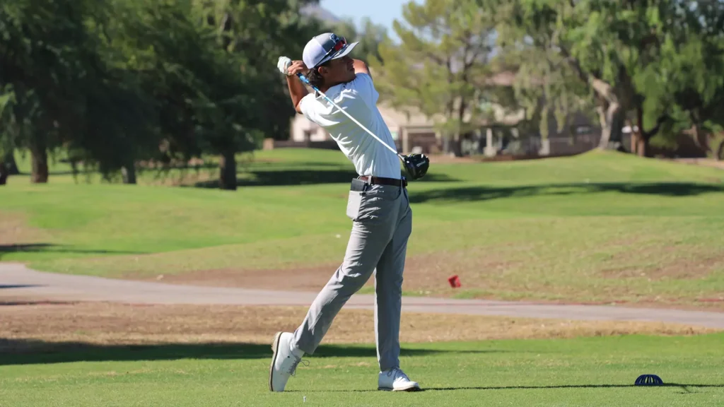 Golfer in white shirt and gray pants mid-swing on a sunny golf course with trees in the background.