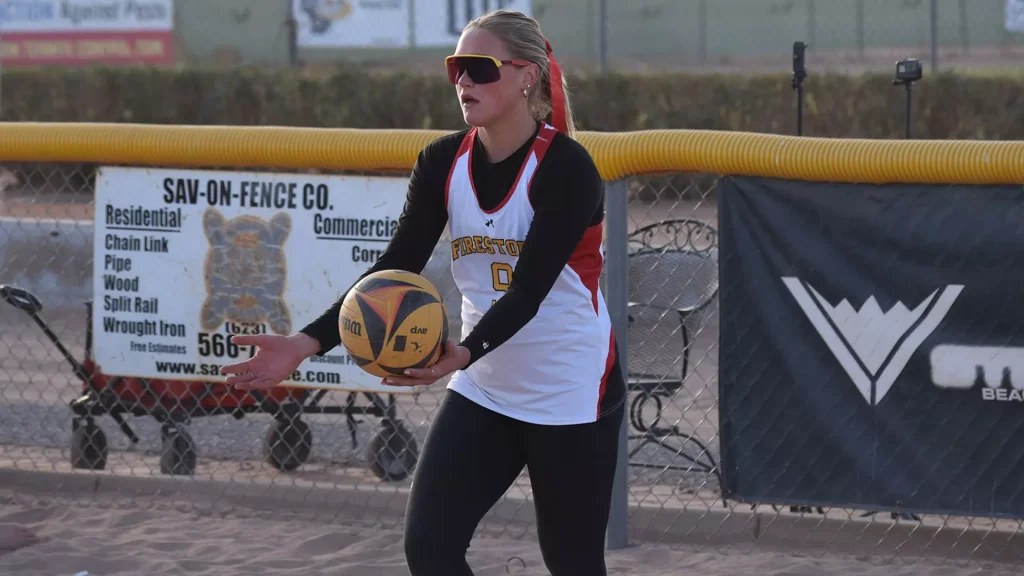 Two Losses At Grand Canyon Classic - Arizona Christian University - Phoenix, AZ Female volleyball player in white and red jersey prepares to pass a yellow volleyball on a court behind a chain-link fence, wearing black long sleeves and red sunglasses.
