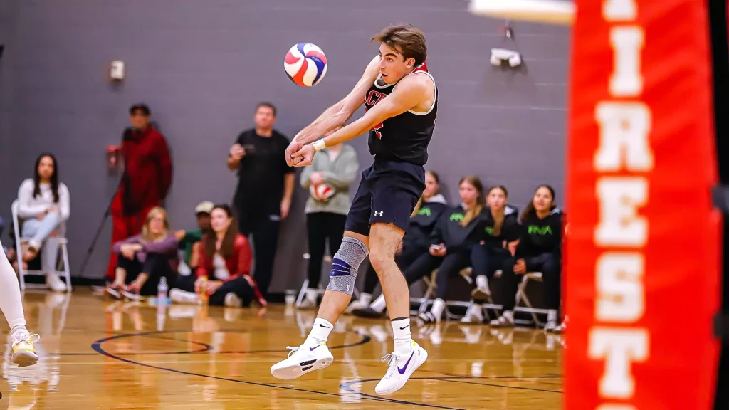 Male volleyball player in mid-air, arms extended to hit a ball during a match in a gym.