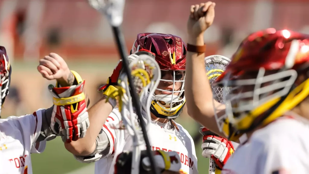 Lacrosse players in white uniforms celebrate on the field with raised arms and sticks in hand, helmeted and joyful.