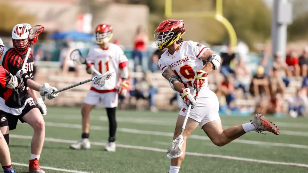 Lacrosse player in white and red Firestorm jersey #9 charges forward with the ball as defenders close in on a sunny field.