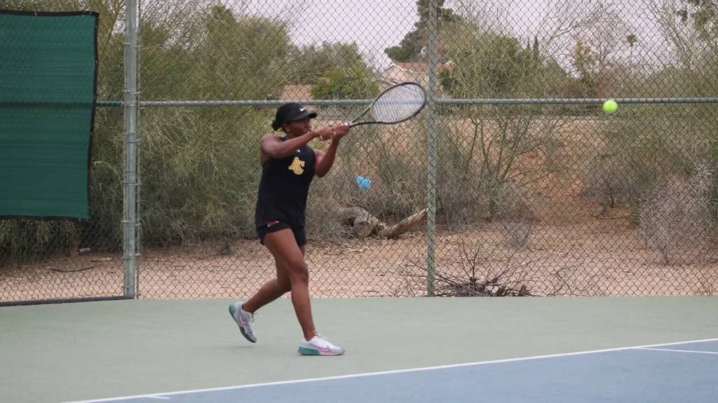 Female tennis player in black athletic wear hits forehand on outdoor court, ball midflight to her right behind a chain-link fence
