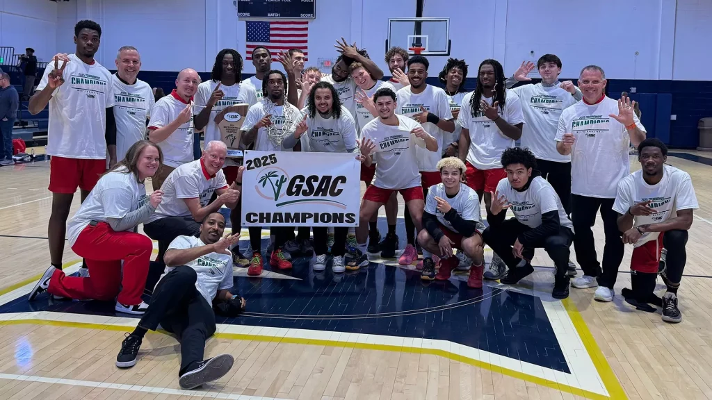 Basketball team posing in a gym holding a GSAC Champions banner and trophy, everyone smiling and making celebratory gestures.