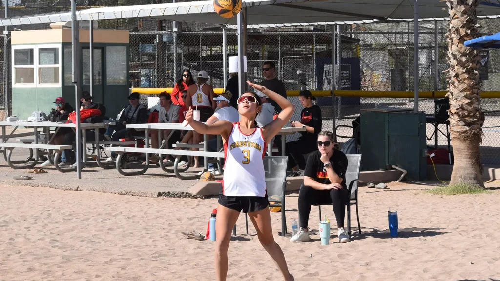 Woman in a white Firestorm jersey with number 3 serves a volleyball on a sandy court, palm trees and bleachers in the background with spectators watching behind a chain-link fence.