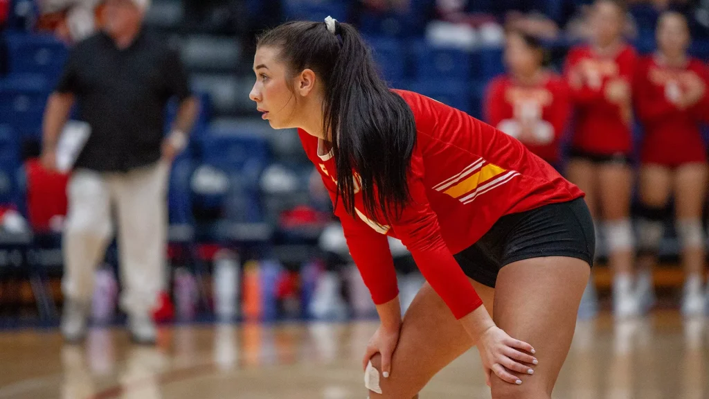 Female volleyball player in a red jersey and black shorts bends over on the court, focused on the play as spectators blur in the background