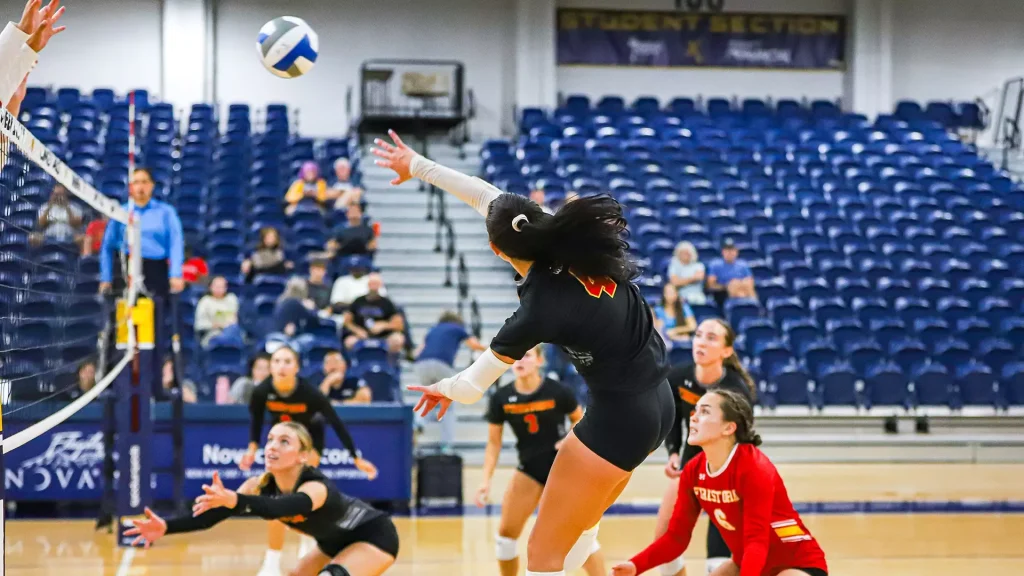 Volleyball action shot: a player in black jumps to spike the ball at the net while teammates dive on the court in a gym with blue bleachers behind.