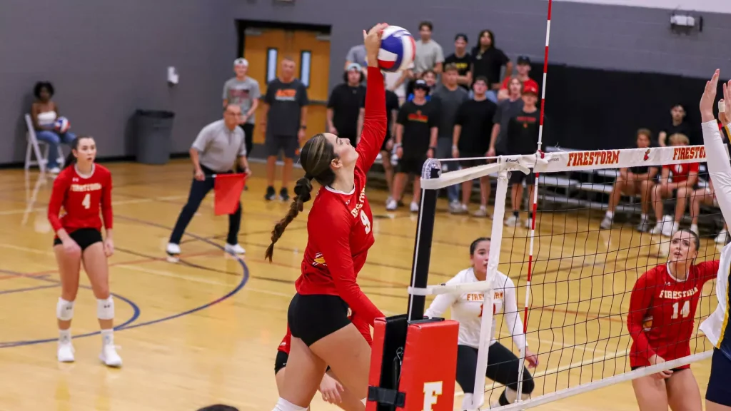 Female volleyball player in red jumps to spike the ball over the net as a crowd watches in a gymnasium.