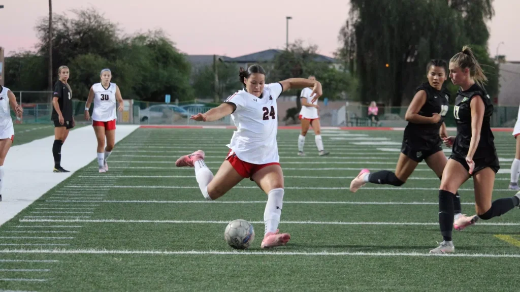 Female soccer player in white jersey number 24 kicking the ball during a match on a green artificial turf field, surrounded by players in black and white uniforms.