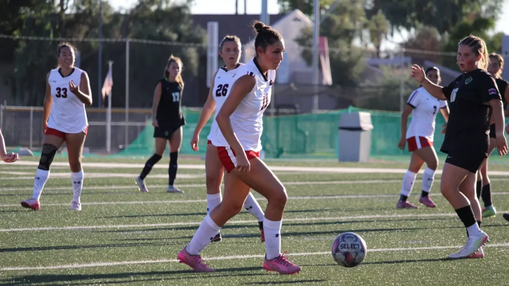 Female soccer players compete on a green turf field; central player in white jersey and red shorts dribbles the ball with teammates around.