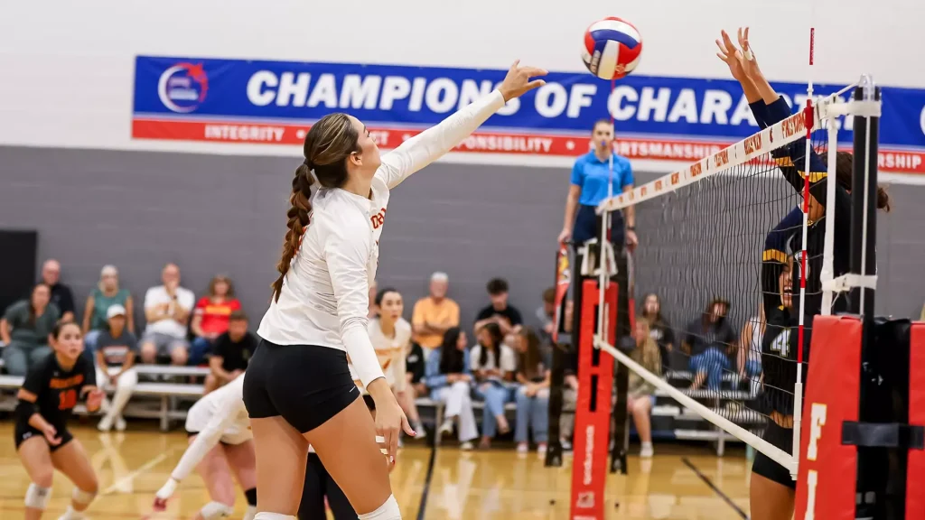 Female volleyball player in a white jersey reaches to spike the ball over the net as teammates and spectators watch in a gymnasium.