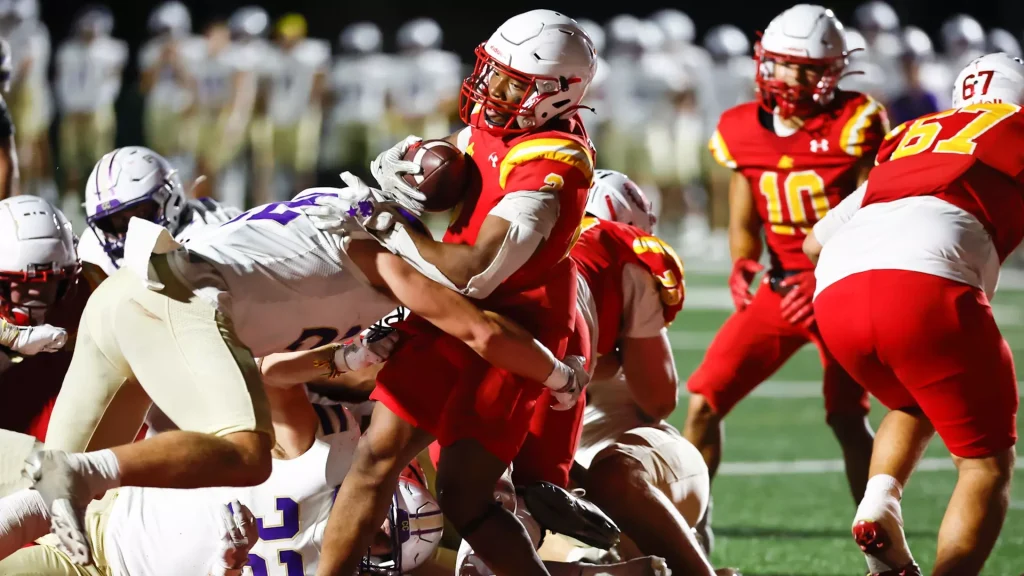 American football game action: a player in red holding the ball is tackled by multiple defenders in white uniforms at night on a green field.
