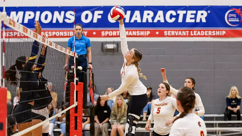 Volleyball player in white spikes the ball over the net as teammates watch from the court in a gym with a 'Champions of Character' banner behind them.