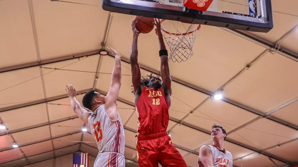 Basketball player in a red uniform (number 12) dunks the ball over two white-uniform opponents inside a gym, with a defender watching nearby.