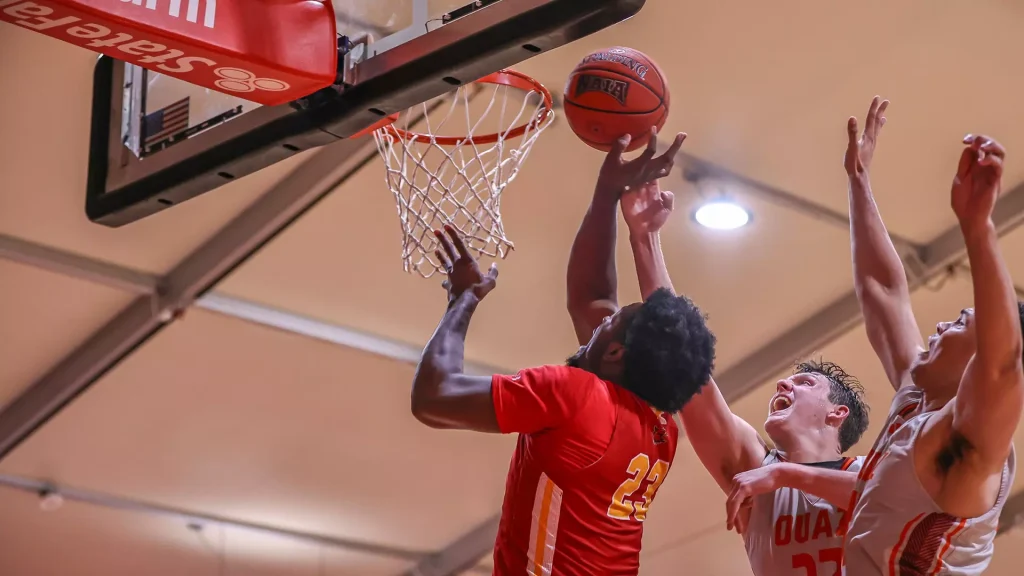 Two basketball players in red and white jump for the ball at the hoop during a game, with the net and backboard visible above them.