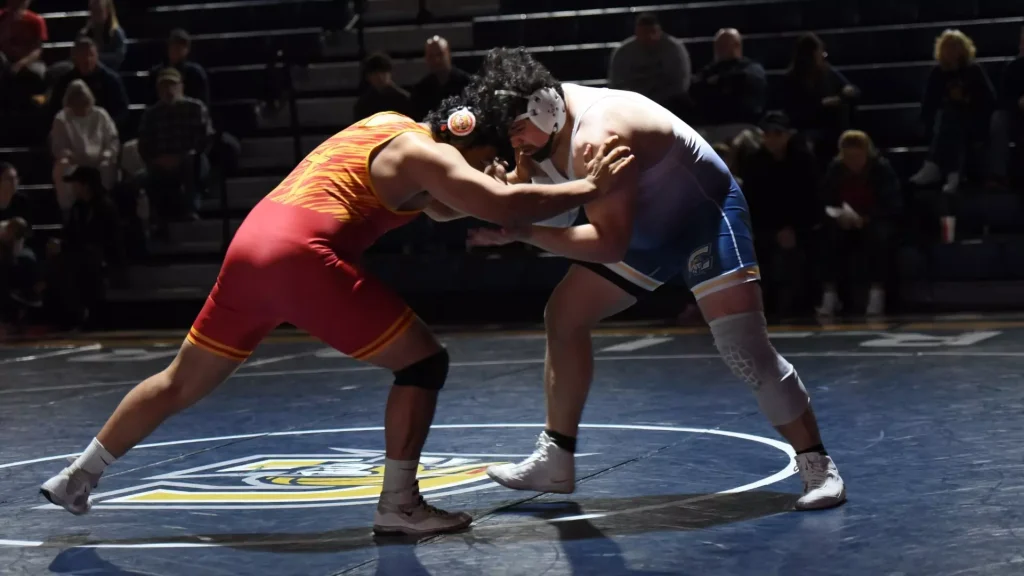 Two male wrestlers in red and blue singlets grapple in a face-to-face clinch on a blue wrestling mat, with spectators in the stands behind them.