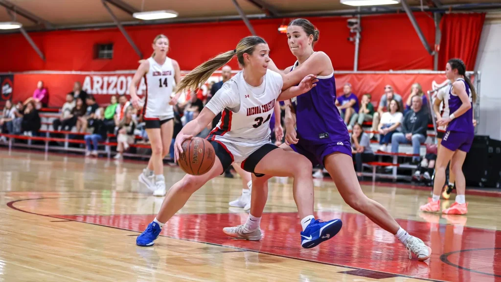White-jersey player dribbles fast toward the basket while guarded by a purple-jersey defender on a polished gym floor with spectators in the stands behind them.