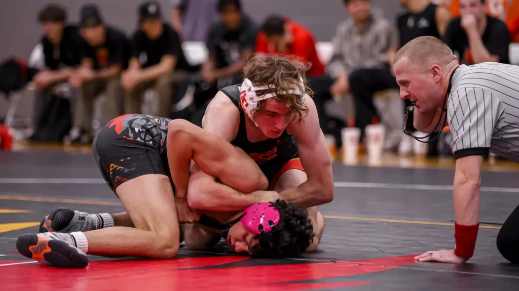 Two wrestlers grapple on a black mat as a referee watches from the right and a blurred crowd looks on in the background.