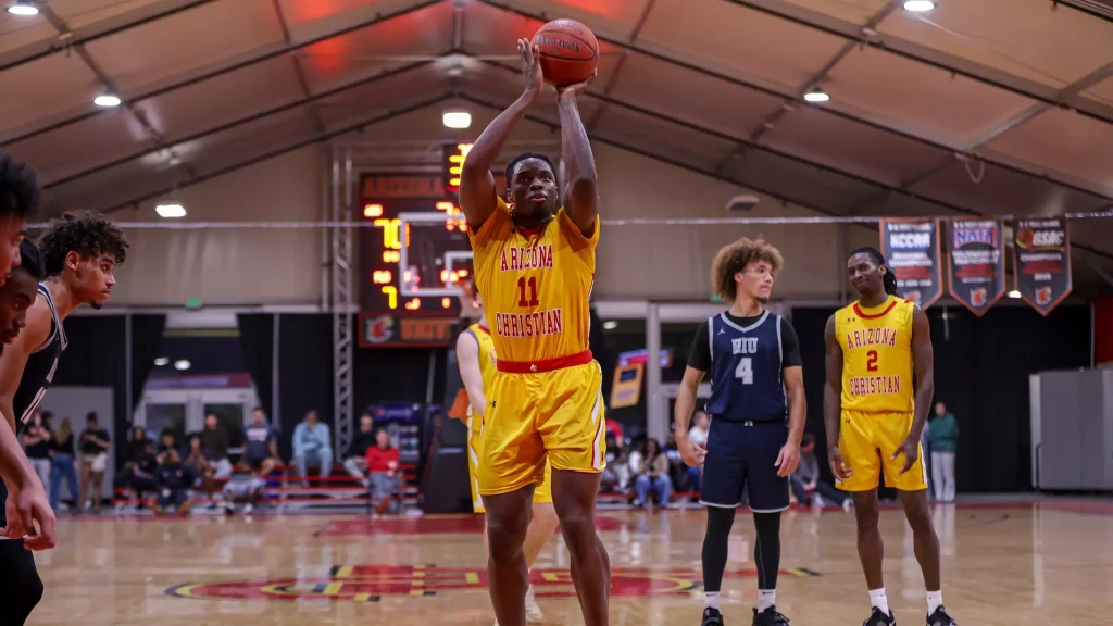 Arizona Christian player in yellow jersey (#11) shoots a jump shot while teammates watch in a gymnasium.