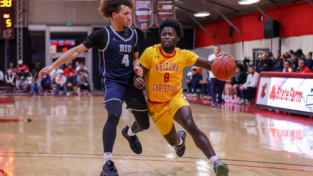 Basketball player in a yellow Arizona Christian uniform dribbles the ball while a defender in navy blue pursues on a polished gym floor.