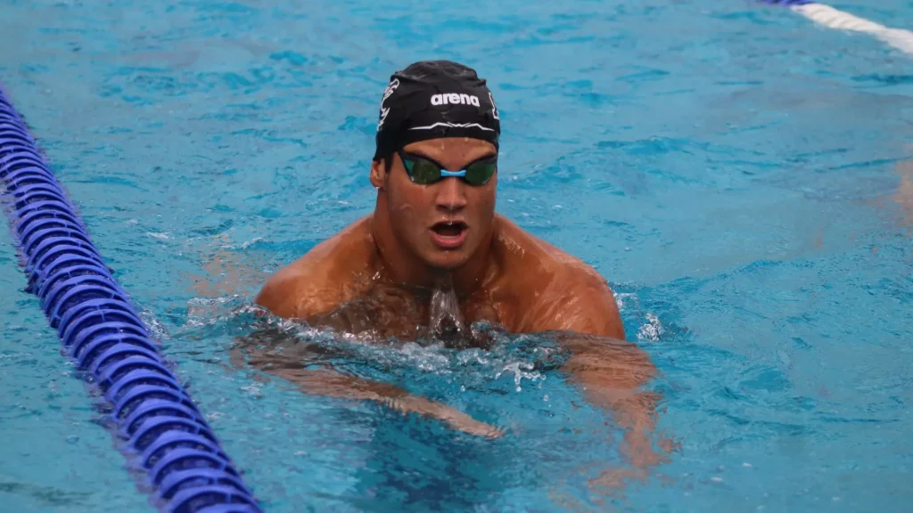 Male swimmer wearing a black Arena cap and green goggles surfaces in a blue pool beside a lane rope, mouth open as he breathes.