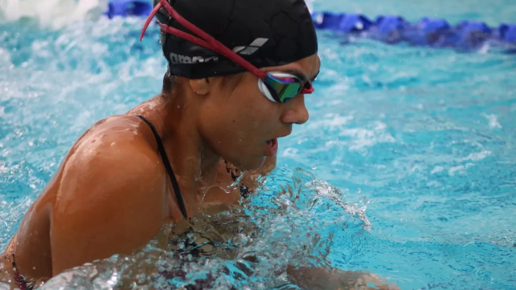 Swimmer in a black swim cap and mirrored goggles paddles through turquoise pool water, breathing near the surface.