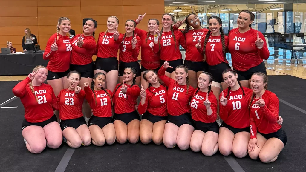 Group of female volleyball players in red ACU jerseys pose together in a gym, smiling and raising one finger in celebration.