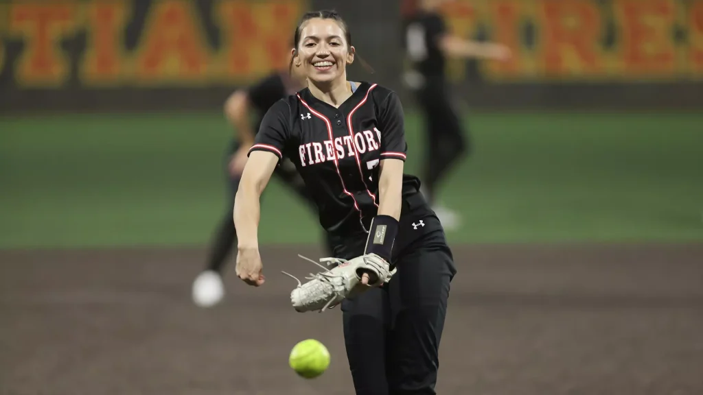 Smiling female softball pitcher in a black Firestorm uniform throws a pitch on a dirt infield; a bright green ball is in the foreground.