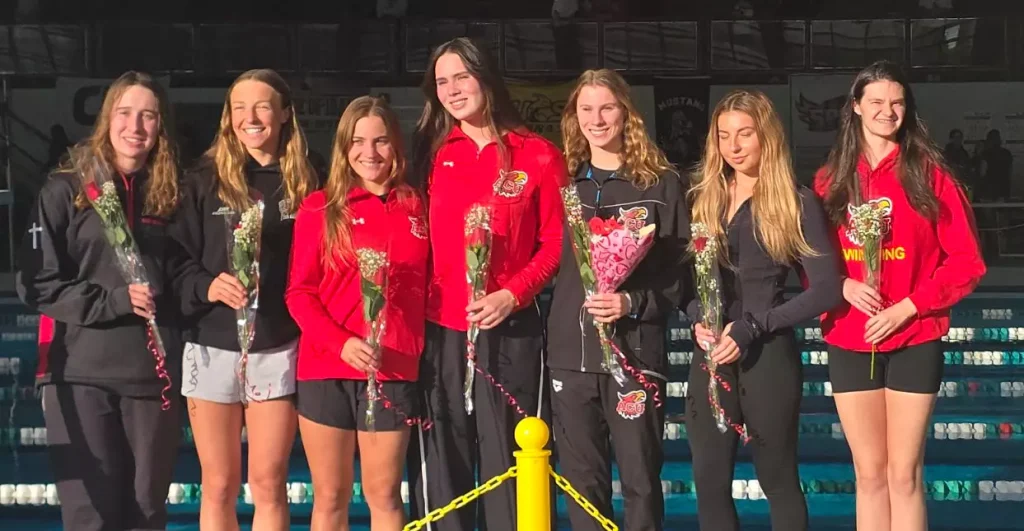 Seven young swimmers stand by an indoor pool, each holding a bouquet of flowers and smiling.