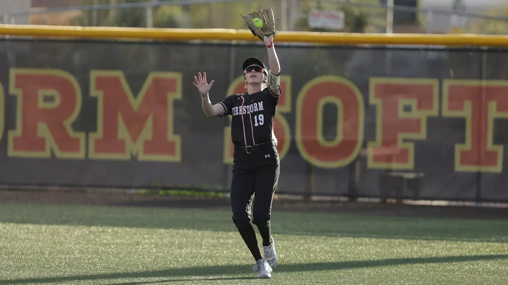 Female softball player in black uniform reaching up with glove to catch a ball on the field; background shows a large red and yellow letters fence.