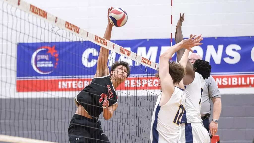 Volleyball players jump at the net during a match; a player in black spikes the ball while teammates reach up.
