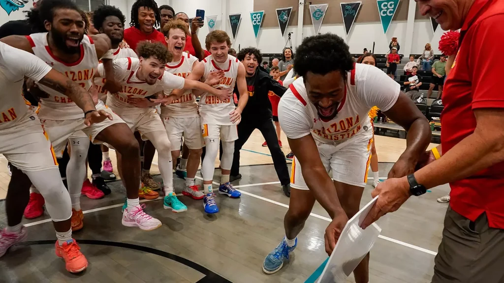 Arizona Christian basketball players in white uniforms celebrate in a gym, huddled together in a joyous group with fans watching in the stands.