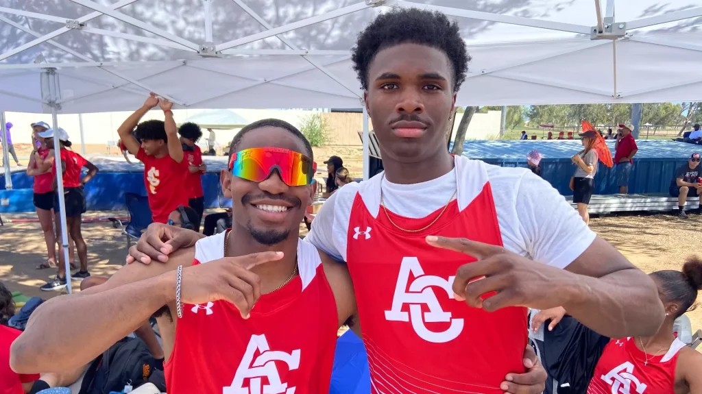 Two young men in red athletic jerseys pose for a selfie under a white canopy at an outdoor event, smiling and showing peace signs.