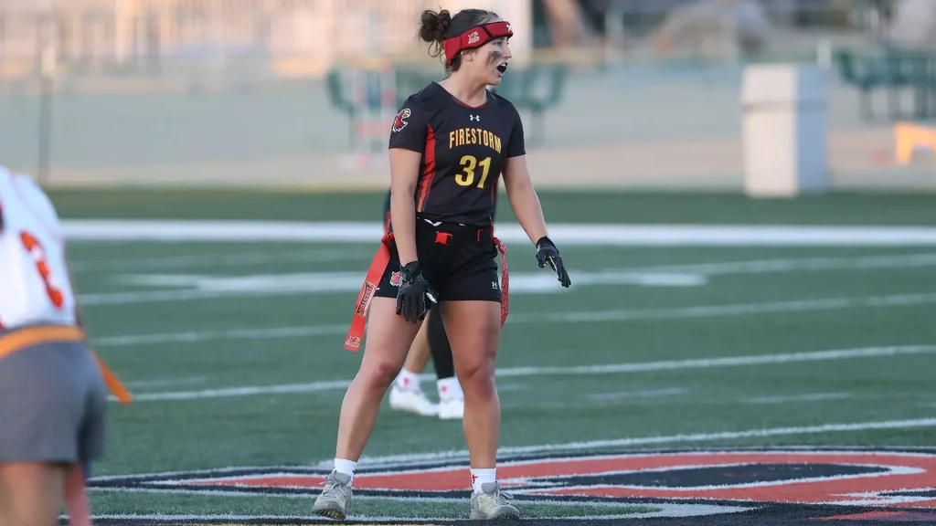 Female lacrosse player in a black and red Firestorm uniform #31 stands on the turf, shouting or signaling during a game with gloves and a red headband.